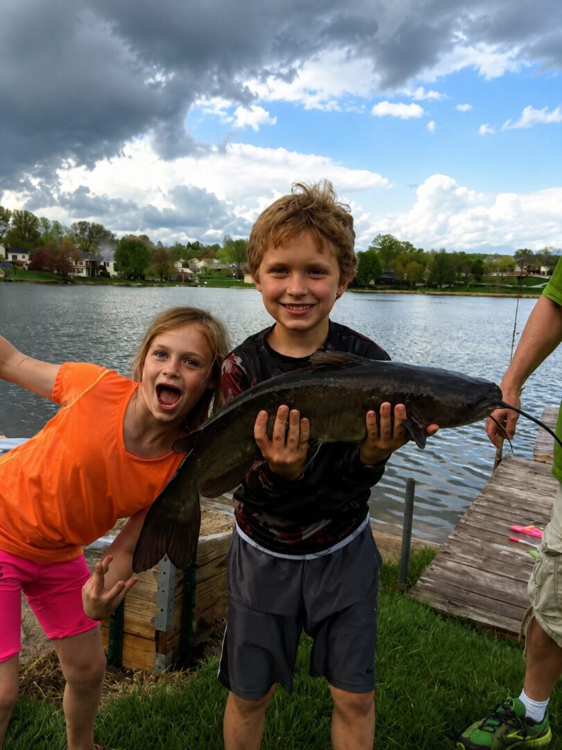 Kids proudly holding a large catfish by lake.