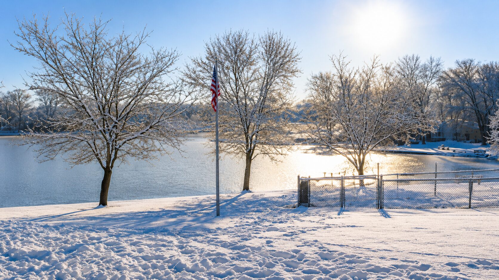 Snowy lakeside with trees and flagpole.